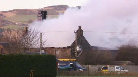Smoke billows from the remains of a single-storey stone house, which no longer has a roof. In the foreground there is a hedge and a couple of vans, and there are hills and a tower in the background.
