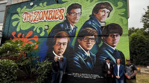 The Rocks Management A mural depicting members of the music group The Zombies on the side of a building with members of the band standing in front of it. The mural is light green and dark green and depicts five men wearing blue velour or velvet jackets. They have 60s style hair and the text is written in a 1960s-style font.