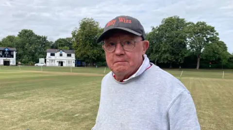 A man wearing glasses and a dark grey cap that has the words 'Billy Wizz' spelled out in orange letters above the peak. Behind him there are two white-washed cricket pavilions and a yellow/green grass cricket pitch.