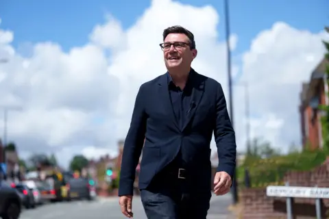 AP Andy Burnham smiles during a campaign visit to Ashton-in-Makerfield before the forthcoming by-election, in Manchester, England, Tuesday, June 9, 2026. He is wearing a dark suit jacket with a dark shirt underneath. He is smiling and waling in a sunny street. His hair is dark and he is wearing dark-framed glasses.