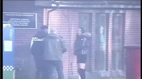 Greater Manchester Police  Stephanie Heaps stands outside prison wearing over the knee black heeled boots, a black skirt and a coat with furry hood, with two men seen to the left of her, their faces not visible