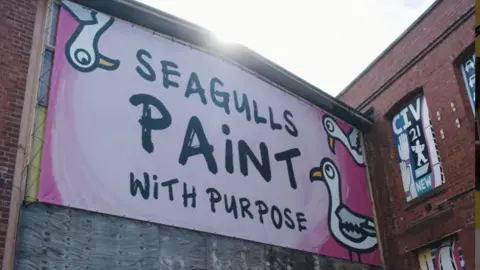 Seagulls A large pink banner sign for Seagulls Paint - it says Seagulls - paint with purpose and has three cartoon drawings of seagulls on. 