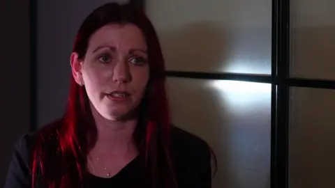 Sarah Myatt, who has dyed red hair and is wearing a black top with a necklace, speaks to he camera in an office with a perspex screen in the background