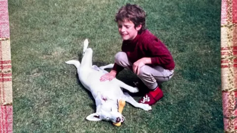 Sue Neale A grainy film photograph taken in the 1980s of a young boy playing on the grass with a white dog, who has a rubber bone in its mouth. The boy has short curly dirty blond hair and is wearing a red jumper, red slippers, and grey jogging bottoms. His eyes are closed and he is smiling.