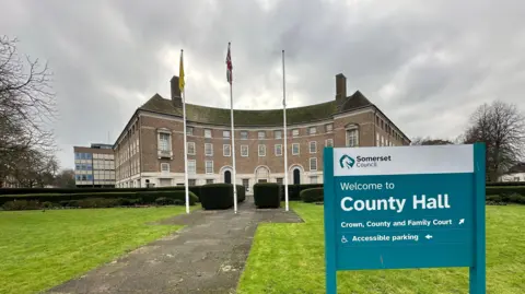 BBC A large brown four-storey building with Georgian-style windows and three flag poles outside. There's a blue sign on the green grass at the front saying 