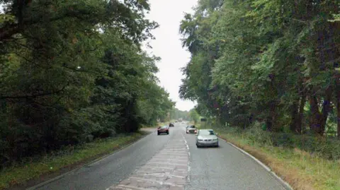 A Google street view image of a single carriageway main road in the countryside. Lots of trees are either side of the road and cars are travelling in both directions.