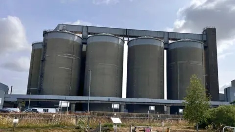 Martin Giles/BBC A factory site with large metal silos against a blue sky on a sunny day.