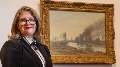 Blackpool Council Paulette Brien who has long brown hair and large spectacles is wearing a white shirt with black stripes, a black bow and a black velvet jacket.
She stands in front of the Monet.