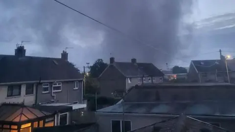 Siân Thomas Residential houses in front of a large cloud of black smoke rising in the sky. 