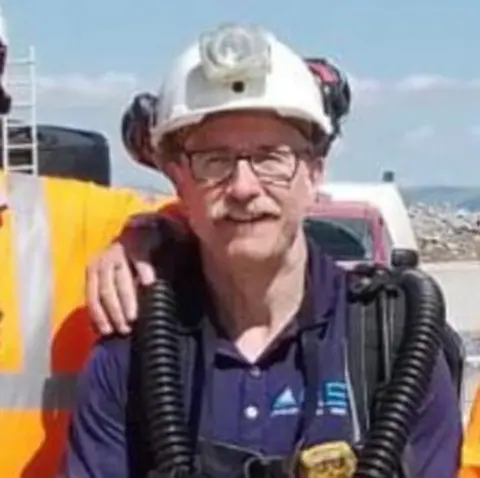 Caroline Thwaites Colin Thwaites wearing a white hard hat with a lamp fitted to the front. He is standing outdoors and it is a sunny day.
