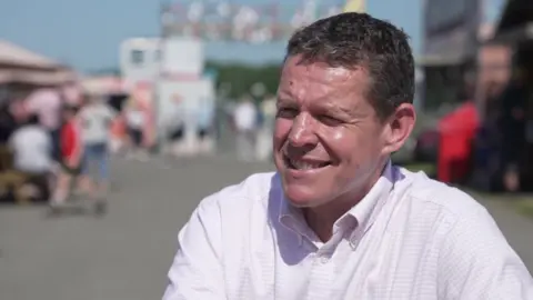 Rhun ap Iorwerth smiling to a camera, wearing a pink shirt and speaking with a festival in the background