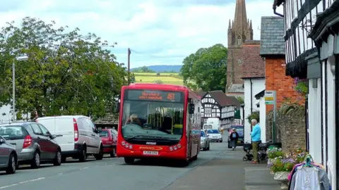 A red single-decker bus drives up a steep road. It says "Hereford via Lyonsall" on the front, plus the number 461. There is a row of shops and a church on the right side of the street.