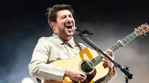 Getty Images Marcus Mumford, wearing a light cream shirt, holding a guitar with his mouth open and a black and silver microphone in front of him