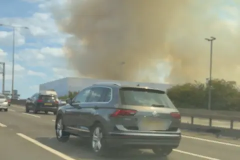 Supplied A silver family car is moving on a motorway. Other cars are in the frame, and in the background, large plumes of smoke rise from an industrial warehouse building.