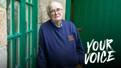 BBC An older man with grey receding hair and glasses leans against a granite wall looking towards the camera. He wears khaki, flannel trousers and a blue sweatshirt. He looks sad.