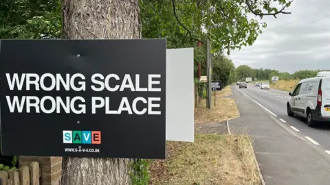 Un cartel negro en un árbol con las palabras en blanco "Escala incorrecta, lugar incorrecto". A la derecha hay una carretera con tráfico.