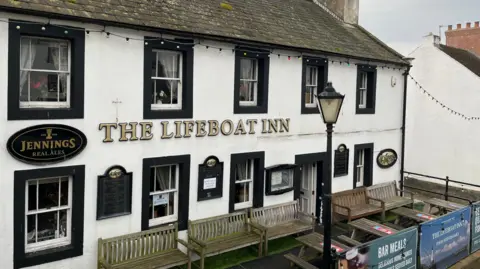 The Lifeboat Inn at Maryport. The two-storey white building has black details around the sash windows and door. The name is on the front of the building in golden lettering. There are wooden benches and picnic tables at the front of the building.