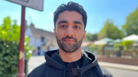 Nicola Haseler/BBC Aman Thakkar standing outside by a sign. He has short hair and a beard, and is wearing a black jumper. He is looking straight at the camera and buildings are behind him, but blurred. 