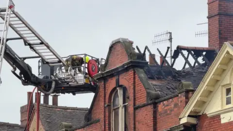 A firefighter working at the top of a building on an aerial platform. The roof supports of the building are charred and a chunk of the roof has fallen away. There are chimneys and aerials alongside the section of roof.