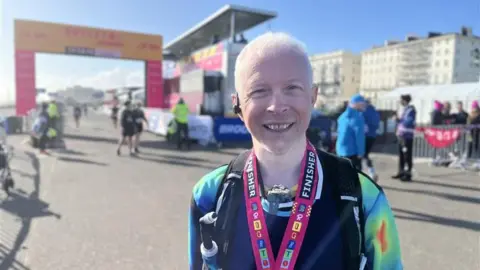 Clarke Reynolds in running kit with a medal around his neck. A race finish line can be seen in the background and he is smiling at the camera.