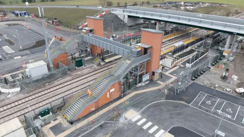 An aerial image of Newsham Station with a lift and passenger bridge in the foreground and a road bridge behind and a large carpark on the far side of the line.