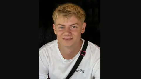 Via Devon and Cornwall Police Fergus Lewis smiles at the camera. He has blond hair and blue eyes and wears a white Billabong branded T-shirt and has the strap of a bag over his shoulders. 