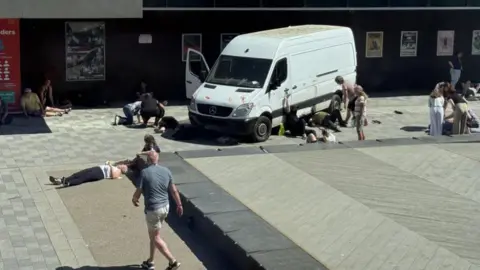 Grace McGrory/BBC A white van with red hand marks on it and several people walking, standing around or lying on the floor. You can see someone holding on to the side of the van and someone else lying underneath it in front of a building with several posters outside.