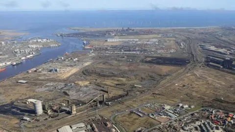 Teesworks An aerial view of the Teesworks site. The mainly undeveloped industrial land sits at the mouth of the River Tees as it enters the North Sea. Buildings are dotted around the land. There are dozens of wind turbines just off the coast.