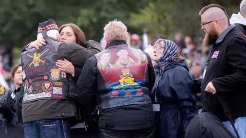 National Memorial Arboretum A group of people at Ride to the Wall. A woman is hugging a man who has badges on the back of his jacket, one which says 'Army' with an emblem above it. Next to him an attendee has a jacket with the faces of soliders on it. 