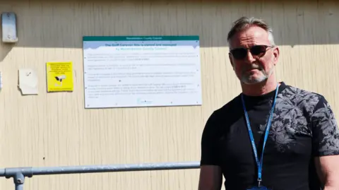 A man in a black T-shirt standing outside a wood clad building. He has dark hair greying at the sides and is wearing a badge on a blue lanyard and dark glasses.