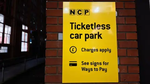 A bright yellow NCP sign outside of an NCP car park. It says in black letters: "Ticketless car park. Charges apply. See signs for ways to pay."