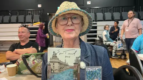 A woman wearing a straw hat with glasses on. She is wearing a denim jacket and holding up a diary and a fridge magnet. 