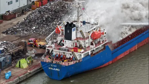 Dale Baxter/BBC A blue and red ship in a dock which has lots of grey smoke billowing out from a fire. The Hull skyline is visible in the distance.