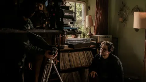 MAB Rabbit Ltd 2024 Director Bryn Chainey sat perched on the floor with his hands on his knees. He sits in front of a busy background full of vinyls, a radio and a lamp either side. To the left is two men stood behind the camera, propping up the equipment. 