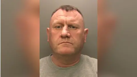 Cumbria Police Paul Irwin's police custody image. He is looking directly into the camera and has short dark hair. He is wearing a grey round-neck top.