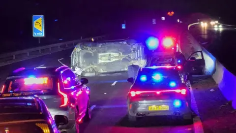 Ken Wheatley Several police vehicles with their blue and red lights on are stopped on the motorway at night. In front of them is a car on its side in the middle of the carriageway.