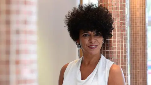 A woman with surly dark hair and a white top stares into the camera