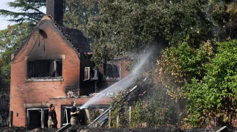 A firefighter sprays water near a burnt-out house