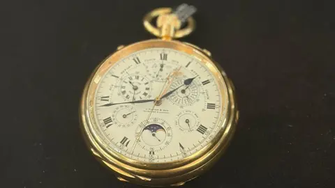 Folleto Una fotografía en primer plano del reloj muestra el reloj sobre un fondo negro. Tiene un borde dorado y una serie de esferas en la esfera.