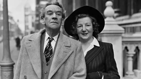 A black and white photo of a Arthur Lucan and Kitty McShane. They are middled-aged and handsomely dressed. Arthur Lucan has short hair and is wearing a blazer coat, cardigan, white shirt and striped tie. To the right, Kitty McShane, with shoulder-length curly brown hair is smiling with lipstick on, a wide-brimmed dark hat and is wearing a stripy, dark coloured blazer jacket and white unbuttoned shirt. A street can be seen behind them.