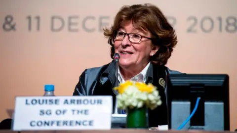 Arbour is seen speaking at press conference in 2018. In front of her is a plaque with her name and her title, and a small pot of flowers. She has short brown hair, and is wearing eyeglasses.
