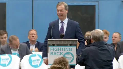 Nigel Farage is photographed as he stands behind a lectern with Brexit Party branding smiling. The placard on the lectern reads Fighting Back. Supporters are seen in Brexit Party t-shirts.
