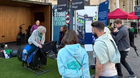 A woman with grey hair and wearing a grey cardigan is riding a horse simulation while people watch on