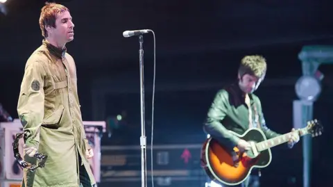 Getty Images Liam and Noel Gallagher stood on stage. Liam is wearing a green Parka coat and holding a tambourine, Noel is playing a guitar.