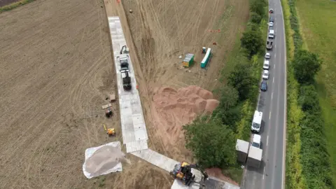 Network Rail An aerial shot of a field with a metal track next to a road, with vehicles parked along the road. There is sand in the field. There are trees and bushes along the road.