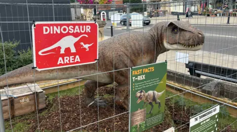 A model of a T-Rex in a cage on a highstreet. The cage has several signs on it which explain more about the dinosaur. 