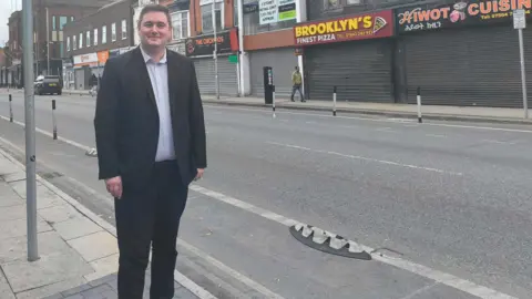 Middlesbrough Council Mayor Chris Cooke, wearing a dark suit, is stood next to a cycle lane with a bin not far behind him. There are shops on the other side of the road.