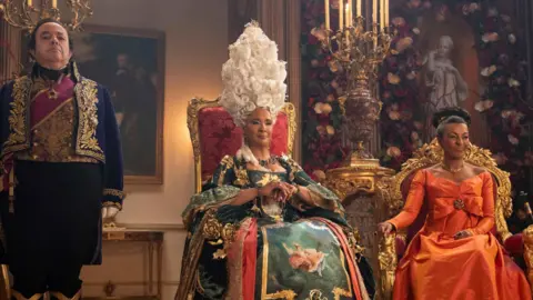 Netflix Queen Charlotte sits in a red and gold throne wearing a tall white wig and green and red dress. She is sat beside Lady Danbury