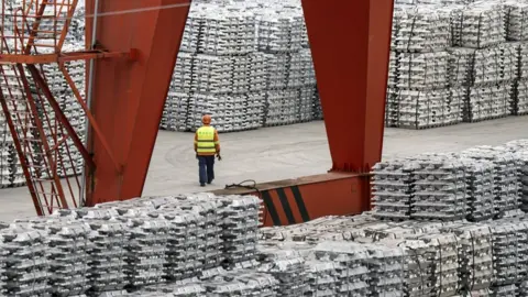 A man walks between stacks of aluminium ingots, stored at a depot in China