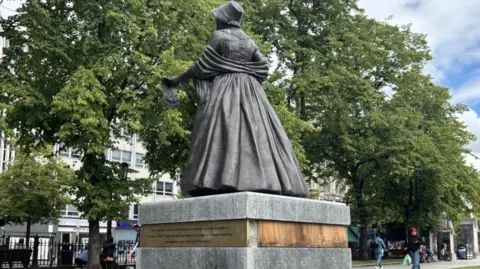 At the base of a statue, a section of plaque is missing, revealing a wooden panel. 